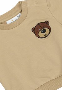 Smėlio spalvos džemperis su apvalia, siuvinėta rudame lape meškos veidu. Ant peties yra du spaustukai, o rankogaliai yra ribotuoti. Minkšta tekstūra.