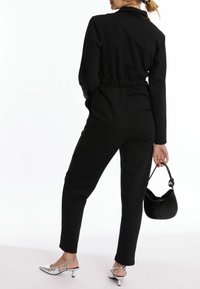 Combinaison noire avec taille cintrée, manches longues et jambes fuselées. Les accessoires comprennent un petit sac noir et des talons métalliques à bride arrière.