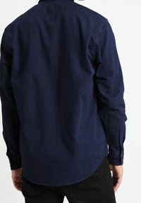 Tmavě modrá džínová košile s dlouhým rukávem, zakončená zaobleným spodním lemem, s manžetami na knoflíky a klasickým límcem. Hladká textura, bez vzorů.