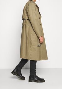 Abrigo trinchera caqui con frente de doble botonadura, cintura ceñida con cinturón y bolsillos laterales, combinado con botas Chelsea negras y jeans negros ajustados.