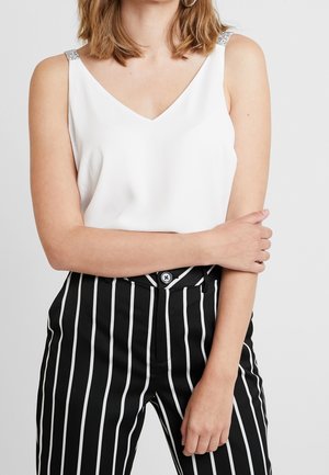 Kobieta w białym, bezrękawnym topie z dekoltem w serek ozdobionym ramiączkami oraz czarnych, wysokowaistych spodniach z pionowymi białymi paskami, z założonymi skrzyżowanymi ramionami.