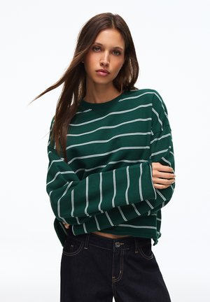 Frau mit langen braunen Haaren, die einen grünen Pullover mit weißen Streifen und dunkle Jeans trägt, stehend mit verschränkten Armen vor weißem Hintergrund.