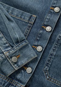 Denimkangas keskmise sinise tooni, hõbedased metallist nupud ja kontrastne pruun õmblus. Omab nupuga klapptaskuid ja tekstuuri viimistlust.