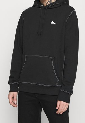 Mężczyzna ubrany w czarną bluzę z kapturem z białymi kontrastowymi przeszyciami i małym białym logo sneakersa na piersi, ręce rozluźnione po bokach.