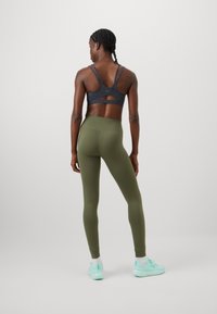 Olíva zöld leggings magas derékkal, fekete sportmelltartóval, amelynek hátul kivágása van. Világoskék sportcipővel viselve.