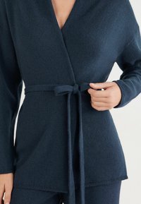 Marineblauer Wickelcardigan aus weichem Strickmaterial, mit V-Ausschnitt, langen Ärmeln und einem gebundenen Gürtel in der Taille für eine figurbetonte Silhouette.