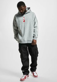 Felpa grigia con cappuccio e un dettaglio grafico centrale rosso, abbinata a pantaloni cargo neri e sneakers rosso e bianche. Tessuto morbido, vestibilità comoda.
