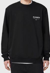 Persona vestida con sudadera negra de manga larga con el texto blanco "ALLSAINTS" y caracteres japoneses en el pecho, combinada con pantalones negros.