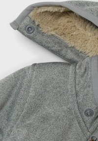 Hoodie en tricot gris avec un extérieur texturé et une doublure en polaire beige douce. Dispose de boutons de fermeture au niveau du cou pour un maintien sécurisé.