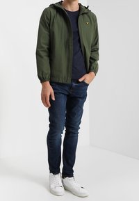 Homme portant une veste à capuche verte, un t-shirt bleu marine, un jean bleu foncé et des baskets blanches, se tenant avec une main dans la poche.