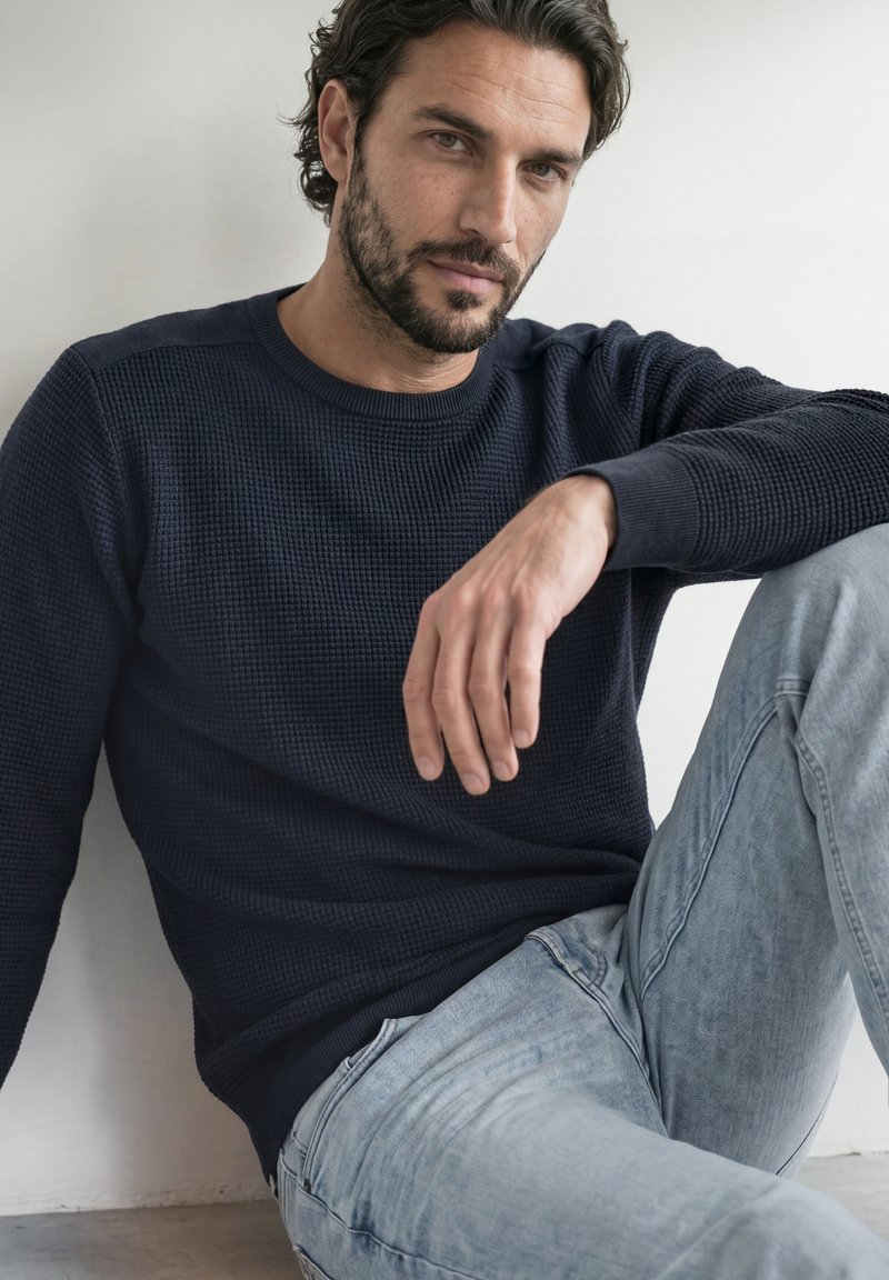 Homme aux cheveux foncés et à la barbe, portant un pull bleu marine et un jean bleu clair, assis contre un mur blanc avec un genou relevé.