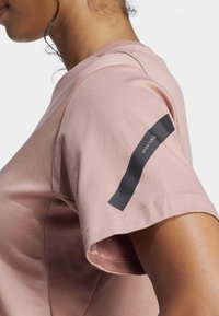 Camiseta corta de manga corta de color rosa claro, hecha de algodón suave. Presenta una franja horizontal negra con el texto "SPORTSWEAR" en la manga izquierda.