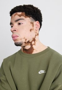 Olivno zelena sweatshirts s krojnim ovratnikom in dolgimi rokavi, ki ima bel Nike logotip na prsih ter mehak, teksturiran material.
