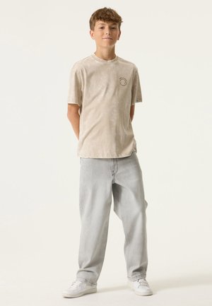 Beige T-shirt met korte mouwen en een circulair logo op de borst, gecombineerd met lichtgrijze wijde jeans en witte sneakers. Soepele textuur.