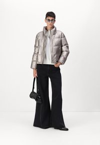 Personne portant une doudoune métallisée argentée, un haut blanc, un pantalon noir à jambes larges, des lunettes de soleil, tenant un petit sac noir, debout sur fond blanc.