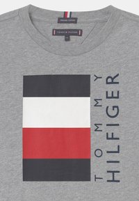 Harmaa orgaaninen puuvillapaita, jossa on suorakulmainen logo tummansinisillä, valkoisilla ja punaisilla raidoilla. Brändin nimi "TOMMY HILFIGER" painettuna tummansinisellä.