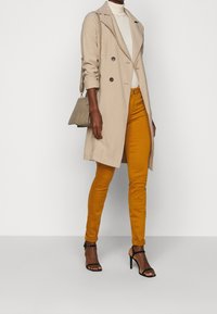 Beige trenchcoat, dubbelknäppt med mörka knappar. Vit polotröja, senapsgula byxor och svarta klacksandaler. Liten texturerad handväska.