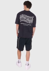 T-shirt oversize noir marine avec un grand texte blanc dans le dos, associé à un short noir avec des finitions blanches, et des baskets blanches.