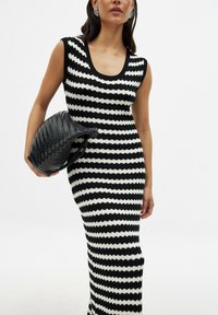 Mustavalkoraidallinen neulemekko, jossa on pyöreä pääntie ja hihaton malli; malli pitää kädessään mustaa punottua clutch-laukkua.