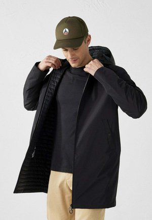 Veste extérieure noire avec fermeture éclair, dotée d'une doublure matelassée. Le modèle porte une chemise noire et une casquette verte.