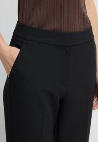Zwarte op maat gemaakte broeken van een gladde stof, met zijzakken en een brede tailleband. Het ontwerp is slank en gestructureerd voor een verzorgde uitstraling.