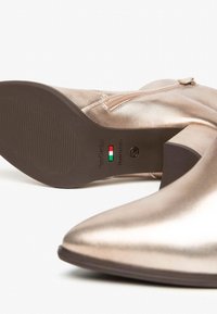 Metallic roségoldene Stiefeletten mit spitzem Absatz, seitlichem Reißverschluss und einer braunen texturierten Gummisohle. Mit Branding auf der Sohle.