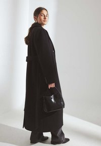 Ilgs melns vilnas mētelis ar pogām un jostu, kombinēts ar melnu somiņu. Sievietes tumšās džinsa bikses un kurpes papildina minimālistisko dizainu.