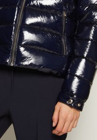 Błyszcząca granatowa pikowana kurtka z srebrnymi zamkami błyskawicznymi i mankietami na zatrzask, noszona na ciemnych dopasowanych spodniach, ręka luźno oparta na boku.