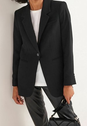 Personne portant un blazer noir sur une chemise blanche et un pantalon en cuir noir, tenant un sac à main en cuir noir sur fond blanc.