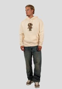 Cream hoodie with graphic of a cartoon figure on the front, paired with blue denim jeans and brown and white sneakers.