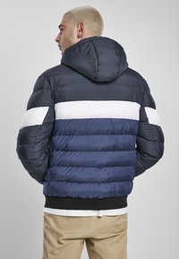 Tumši zila pūkaino jakas ar kapuci ar baltām un gaišāk ziliem horizontāliem svītrām, ribētu apakšmalu un quilted tekstūru.
