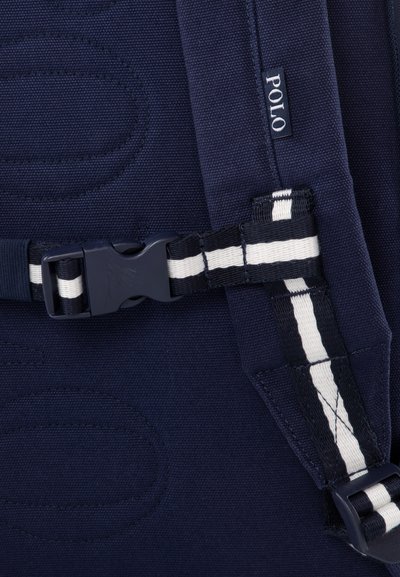 Navyblauwe canvas rugzak met een opvallende gespsluiting met witte accenten en een gestikt logo. Textuurstof met verhoogde patronen.