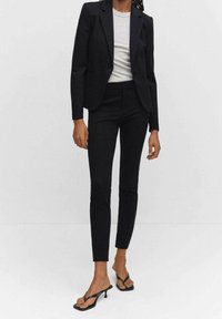 Veste noire cintrée associée à un pantalon slim assorti et un haut gris clair. Des sandales à brides transparentes complètent la tenue.