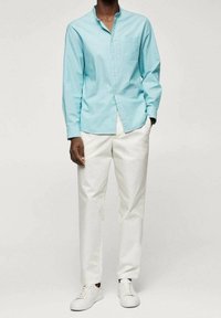 Hellblaues Langarmhemd mit Knopfleiste am Kragen und Brusttasche, kombiniert mit weißer Hose und weißen Sneakers. Glatte Stoffstruktur.