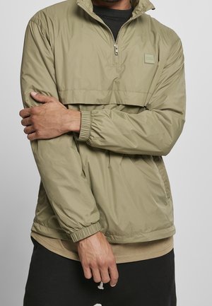 Mężczyzna ubrany w jasnooliwkową wiatrówkę z półzamkiem i elastycznymi mankietami, pozujący z założonymi na siebie na piersi ramionami na jednolitym tle.