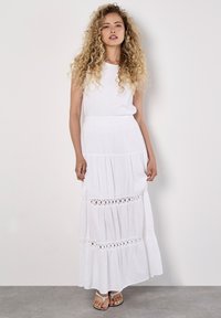 Een model met krullend haar draagt een vloeiende witte maxi-jurk met kantdetails, staande tegen een neutrale achtergrond.