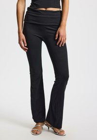 Zwarte flared broek van rekbare stof, met een omgevouwen tailleband. Gecombineerd met zilveren sandalen met straps, wat een strakke silhouet benadrukt.