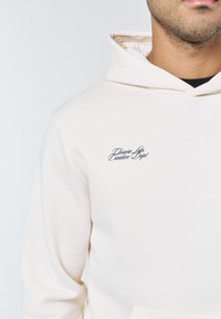 Man iklädd en kremfärgad hoodie med en liten svart broderad text "Closure Ldn Creative Dept" på vänster bröst.