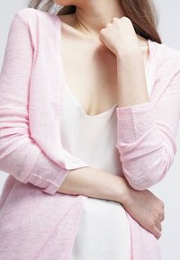 Ljusrosa kofta i ett mjukt, strukturerat stickat material, öppen fram, över en vit topp med rundad halsringning. Långa ärmar är lätt uppvikta.