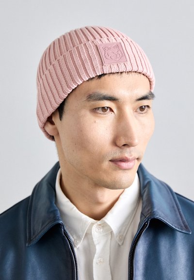 Jonge man met een roze gebreide beanie met een wolflogo, wit overhemd en blauwe leren jas, die iets naar rechts kijkt.
