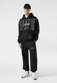 Czarny komplet bluzy z kapturem i spodni dresowych z dużym logo "Champion". Bluza posiada kieszeń z przodu; model nosi białe trampki. Prosty, codzienny design.