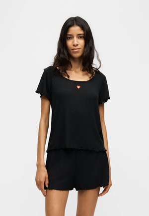 Kvinde med langt mørkt hår iført en sort kortærmet top og matchende sorte shorts med en lille rød hjerte detalje på toppen.