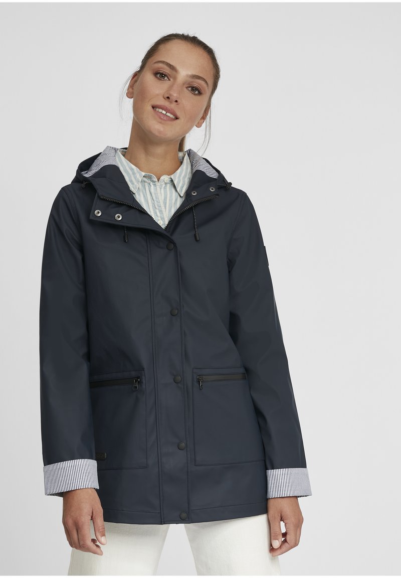 Veste de pluie bleu marine avec une fermeture éclair à l'avant, capuche réglable et poignets rayés. Elle dispose de deux poches zippées et d'une texture lisse et imperméable.