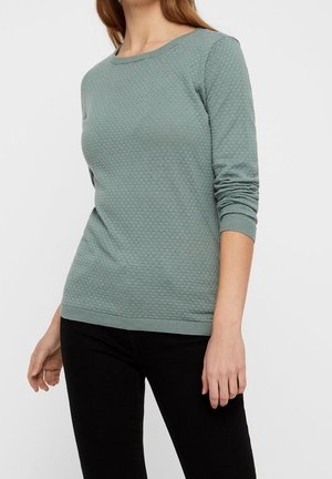 Haut long à manches vert texturé avec un col rond, des poignets côtelés et un ourlet. Porté avec un pantalon noir, présentant un subtil motif en nid d'abeille.