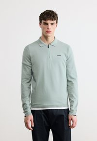 Νεαρός άνδρας με σγουρά μαλλιά φοράει ανοιχτοπράσινη μακρυμάνικη πόλο μπλούζα με ορατό φερμουάρ και λογότυπο "BOSS", συνδυασμένη με μαύρο παντελόνι.