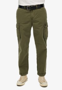 Pantaloni cargo verde oliva con polsini elastici, numerose tasche laterali e zip frontale. Abbinati a una cintura nera e sneakers beige.