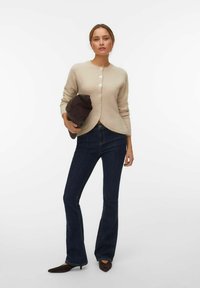 Femme portant un cardigan beige boutonné, un jean évasé foncé et des chaussures pointues foncées, tenant une pochette en daim marron foncé pliée.