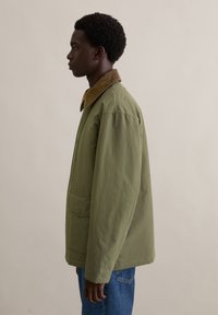 Oliwkowa kurtka z brązowym kołnierzem z sztruksu, przednimi kieszeniami i luźnym krojem, prezentowana na osobie z profilu na neutralnym tle.