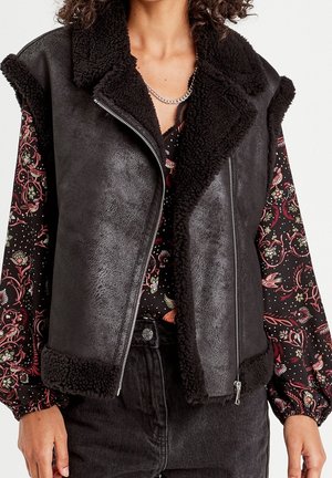 Frau trägt eine schwarze Lammfellweste über einer langärmeligen, floral gemusterten Bluse und schwarzen Jeans, mit einer silbernen Halskette.