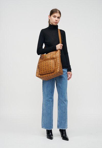 Femme portant un col roulé noir, un jean bleu, des bottes noires, tenant un grand sac à épaule marron à motifs sur un fond clair uni.